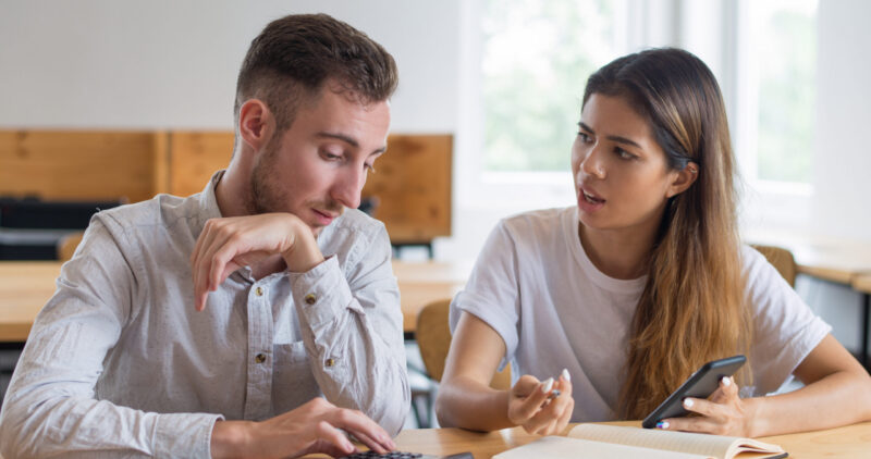 Serious students discussing homework and using digital devices. Young man and woman using calculator and smartphone and sitting at desk in classroom or library. Education and friendship concept.